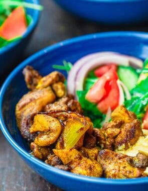 A close up of a chicken shawarma bowl with shawarma chicken, onion, tomatoes, and arugula in a blue bowl against a dark background.
