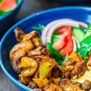 A close up of a chicken shawarma bowl with shawarma chicken, onion, tomatoes, and arugula in a blue bowl against a dark background.