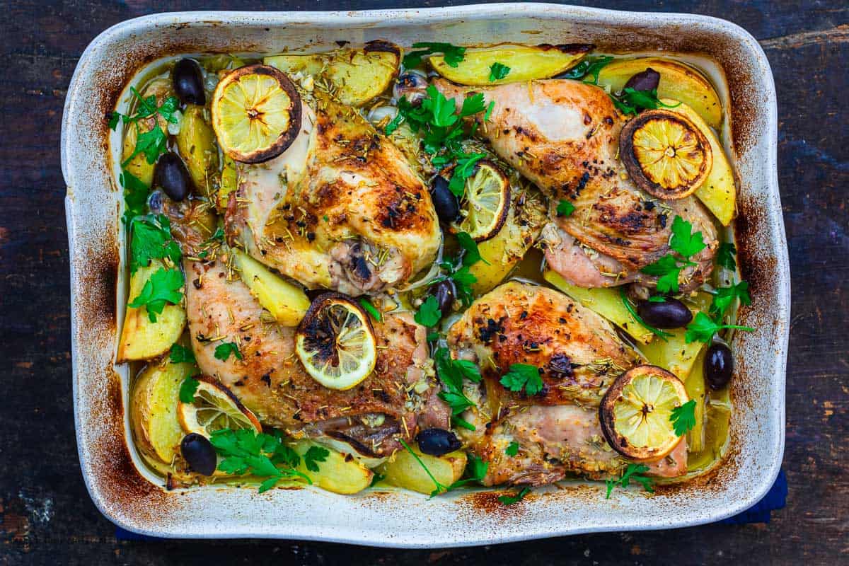 A white casserole dish with Greek chicken and potatoes. Decorated with lemon slices, olives and fresh herbs.