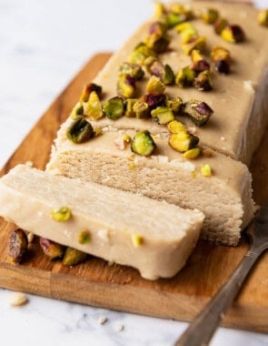 A loaf of halvah topped with roasted pistachios with 2 slices cut on a cutting board with a knife.