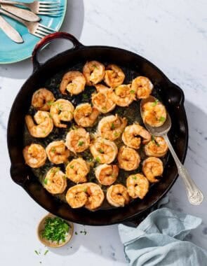 Honey garlic shrimp in a cast iron skillet with a spoon. Next to this is a plate with 2 forks and two knives, a small bowl of chopped parsley and cilantro, and a cloth napkin.