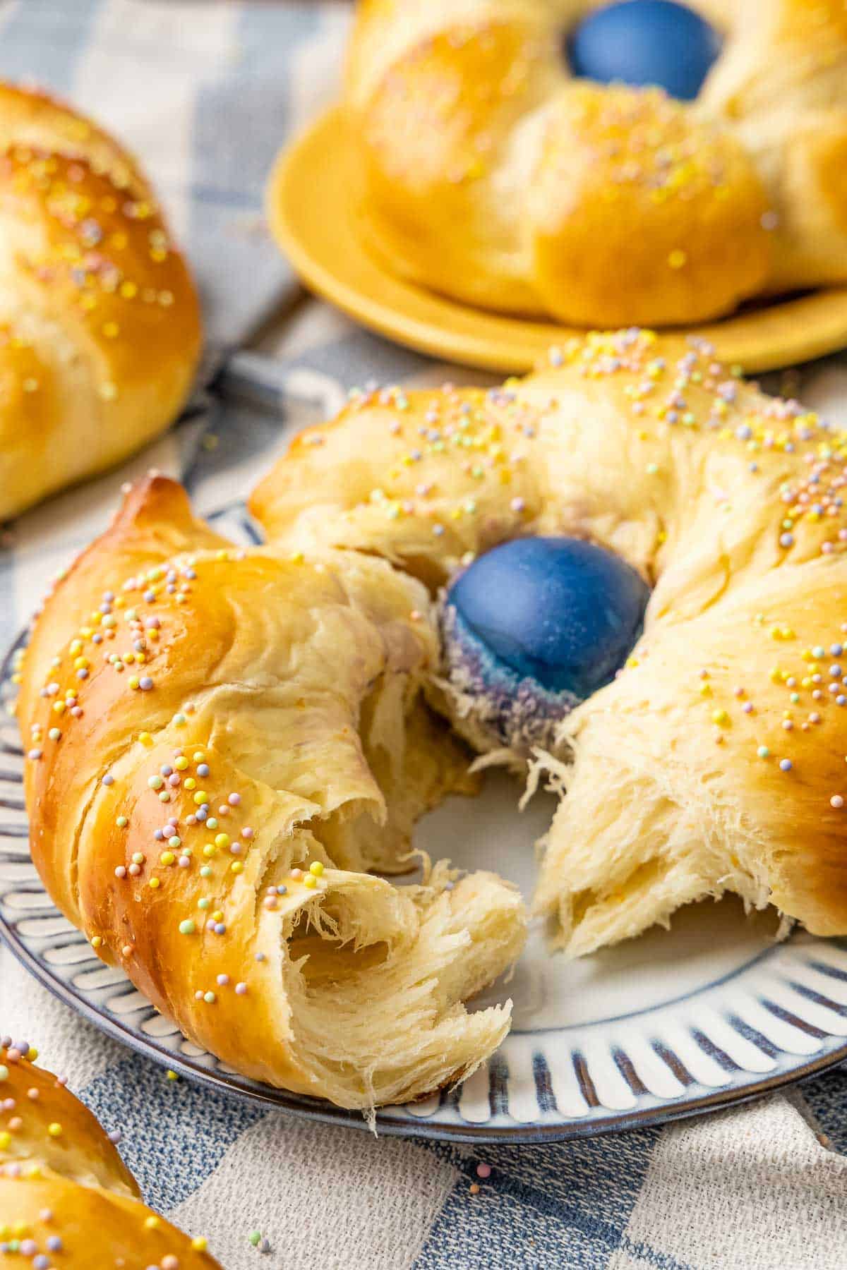 A braided nest of Italian Easter Bread with a naturally dyed blue egg in the center and colored sprinkles. The bread is torn open so you can see the fluffy interior. it's a on white plate with blue edges.