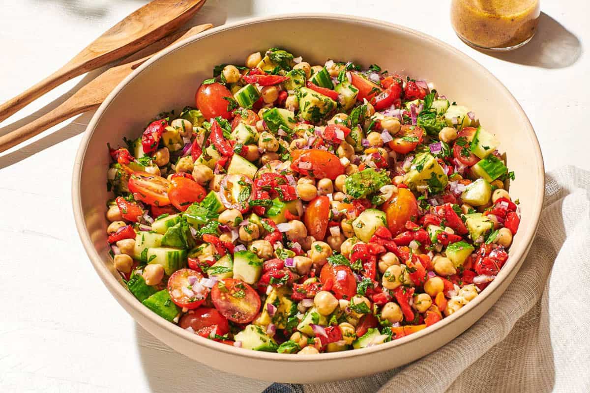 A close up of chickpea salad in a serving bowl next to a wooden serving spoon and fork, a jar of dijon dressing, and a cloth napkin.