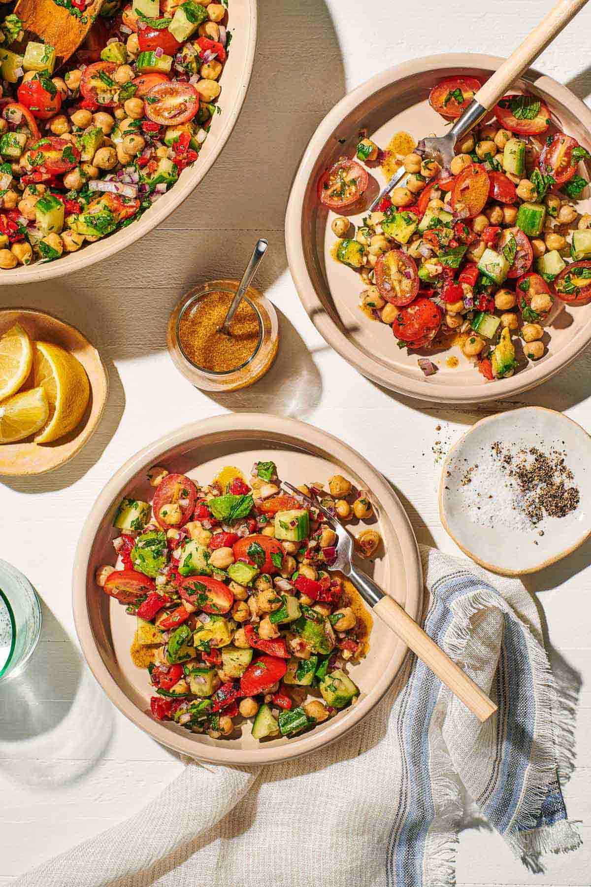 An overhead photo of two bowls of vegan chickpea salad with forks next to a serving bowl of the salad, a small plate of salt and pepper, a small bowl of lemon wedges, a jar of dijon dressing with a spoon, a glass of water and a cloth napkin.