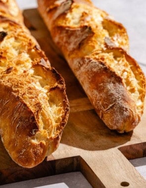 Two baked french baguettes on a wooden serving board next to a cloth kitchen towel and a bowl of jam with a spoon.