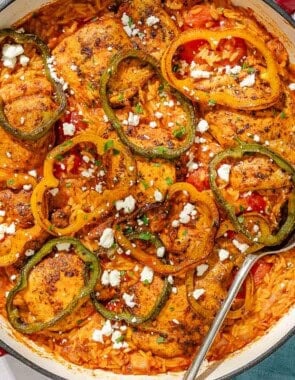 The one pan chicken orzo in a skillet with a serving spoon next to bowls of crumbled feta and chopped parsley, and a cloth napkin.