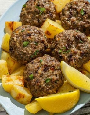 Biftekia with lemon potatoes and lemon wedges on a platter.