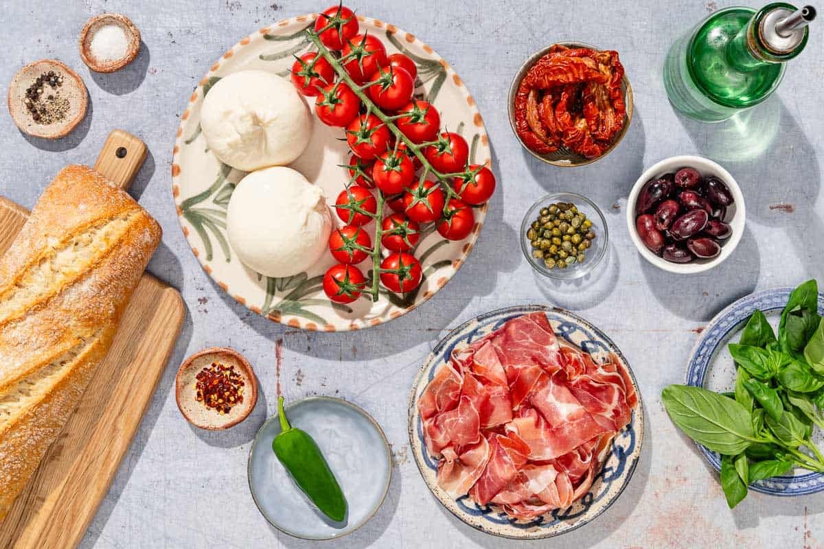 Ingredients for burrata salad including burrata cheese, crusty bread, cherry tomatoes, olive oil, prosciutto, a jalapeño, sun dried tomatoes, kalamata olives, capers, basil, salt, black pepper and red pepper flakes.