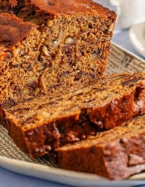 A partially sliced loaf of date nut bread behind 2 slices of the bread on a platter. Next to this is a slice of the bread on a plate.