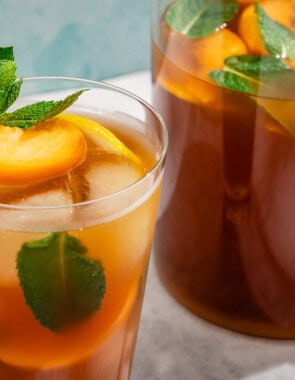 A glass of peach tea garnished with mint and peach and lemon wedges next to a pitcher of the tea.
