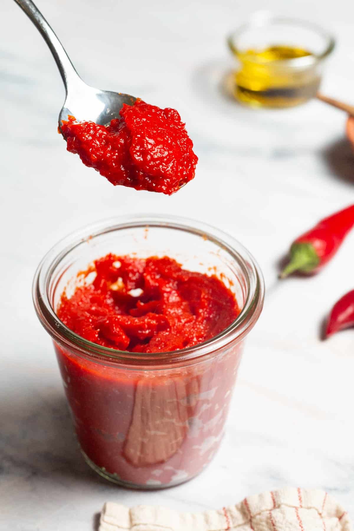 Red pepper paste being lifted out of a jar of the paste on a spoon. Surrounding this is a bowl of olive oil, two red chilies and a kitchen towel.