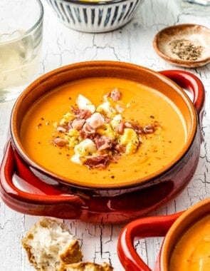 close up of a bowl of salmorejo spanish cold tomato soup topped with chopped hard boiled eggs and prosciutto next to a glass of wine and a bowl of pepper.