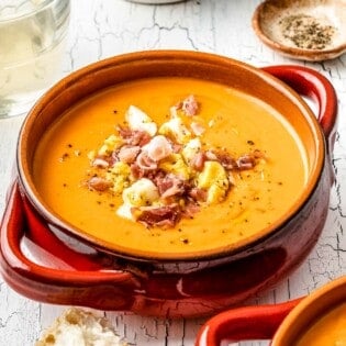 close up of a bowl of salmorejo spanish cold tomato soup topped with chopped hard boiled eggs and prosciutto next to a glass of wine and a bowl of pepper.