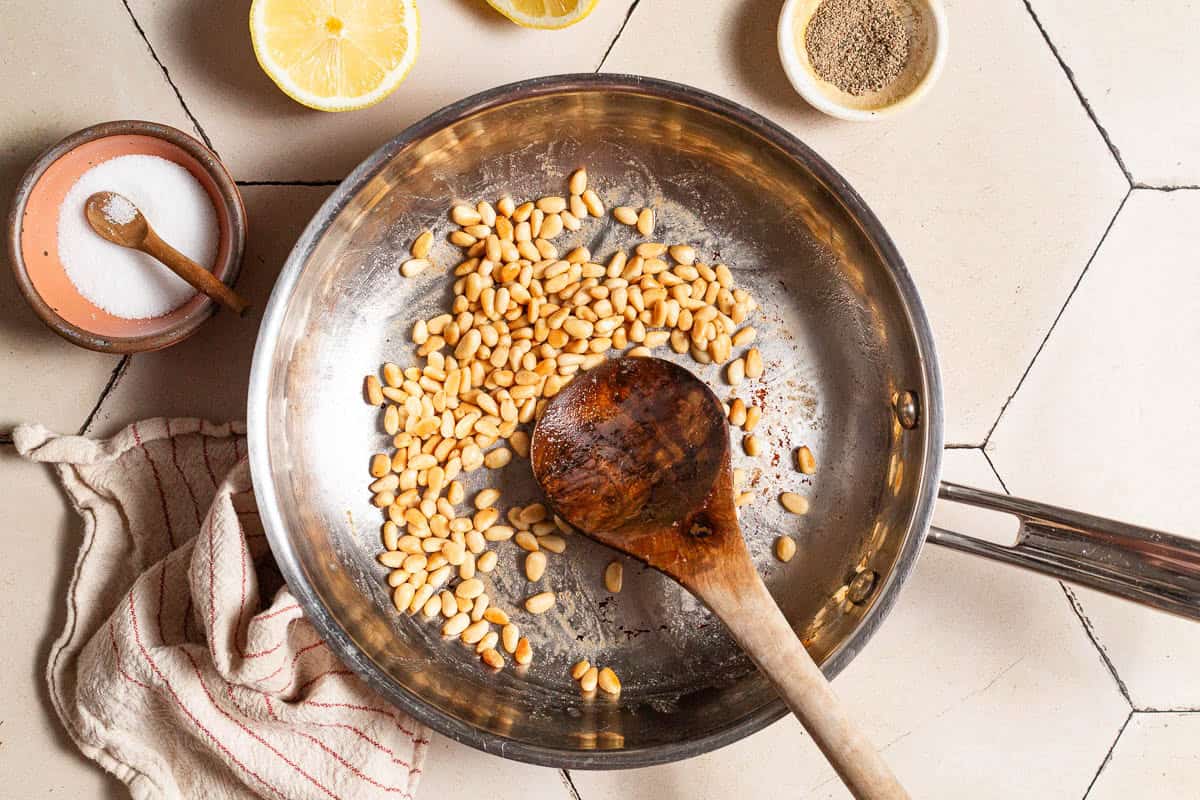 Pine nuts being toasted in a skillet with a wooden spoon. Next to this is a kitchen towel, two lemon halves and bowls of salt, black pepper and pine nuts.