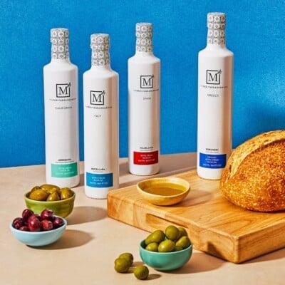 The Everyday Extra Virgin Olive Oil Bundle from The Mediterranean Dish shop next two 3 bowls of olives and a wooden tray with a loaf of bread and a bowl of olive oil.