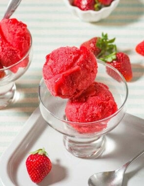 Two scoops of strawberry sorbet in a bowl next to a spoon, several scattered strawberries, a small bowl of strawberries and another bowl of the sorbet.