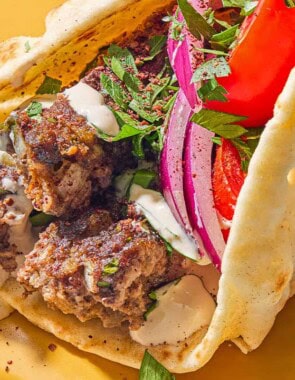 A pita wrapped filled with the sheet pan baked kofta topped with red onion, tomatoes, parsley and tahini on a plate.