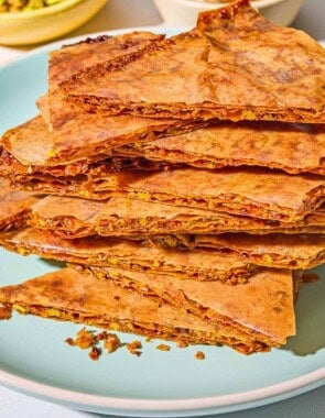 A close up of a stack of baklava bark on a plate. In the background are bowls of chopped pistachios and honey.