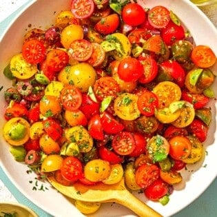 Cherry tomato salad in a serving bowl with a wooden spoon. Next to this is a bowl of chopped parsley, a bottle of olive oil, a bowl of Aleppo pepper and two slices of toasted bread.