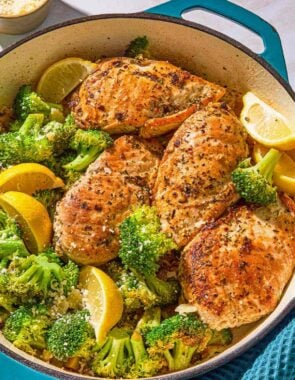 The chicken and broccoli in a skillet with lemon wedges next to a kitchen towel and a bowl of Parmesan cheese.