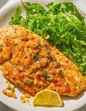 A serving of chicken scaloppine on a plate with an arugula salad and a lemon slice.