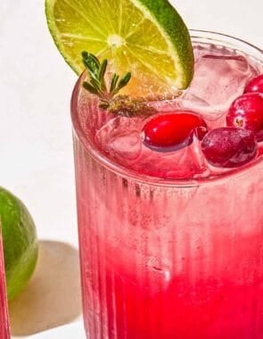 Two glasses of the cranberry mocktail garnished with rosemary, cranberries and lime next to a lime and a bowl of cranberries.
