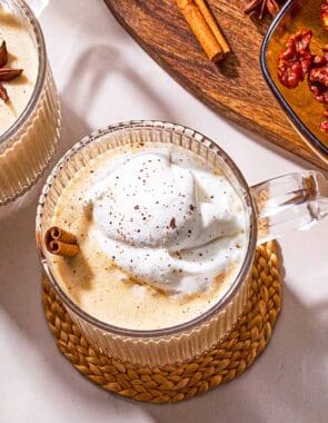 Two mugs of dairy-free eggnog garnished with star anise, a cinnamon stick and whipped coconut cream next to a wooden tray with a bowl of dates, a cinnamon stick a star anise.