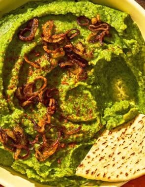 Fava bean dip topped with caramelized onions in a bowl with a pita wedge. This is on a platter with various vegetables and more pita wedges.