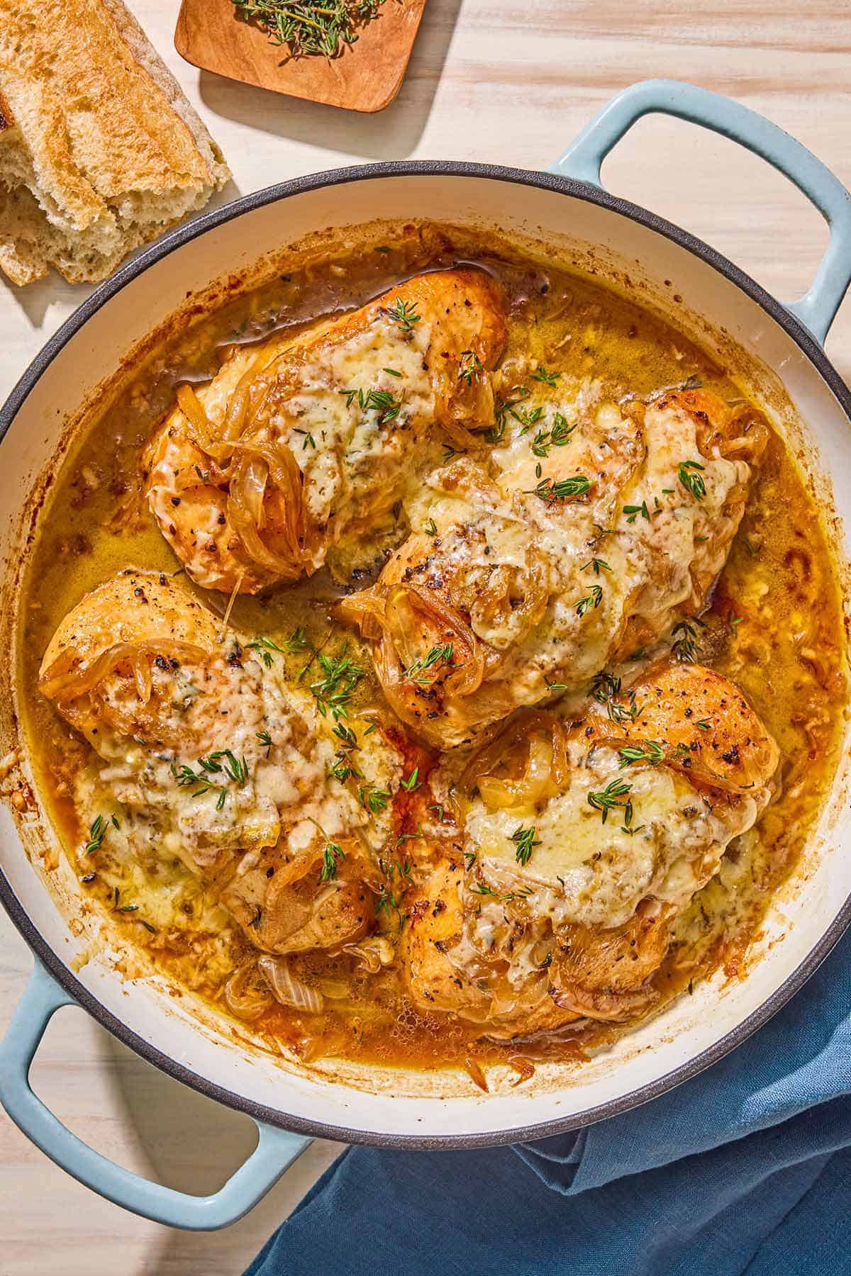 French onion chicken in a skillet next to a bowl of chopped thyme, a piece of crusty bread, and a cloth napkin.