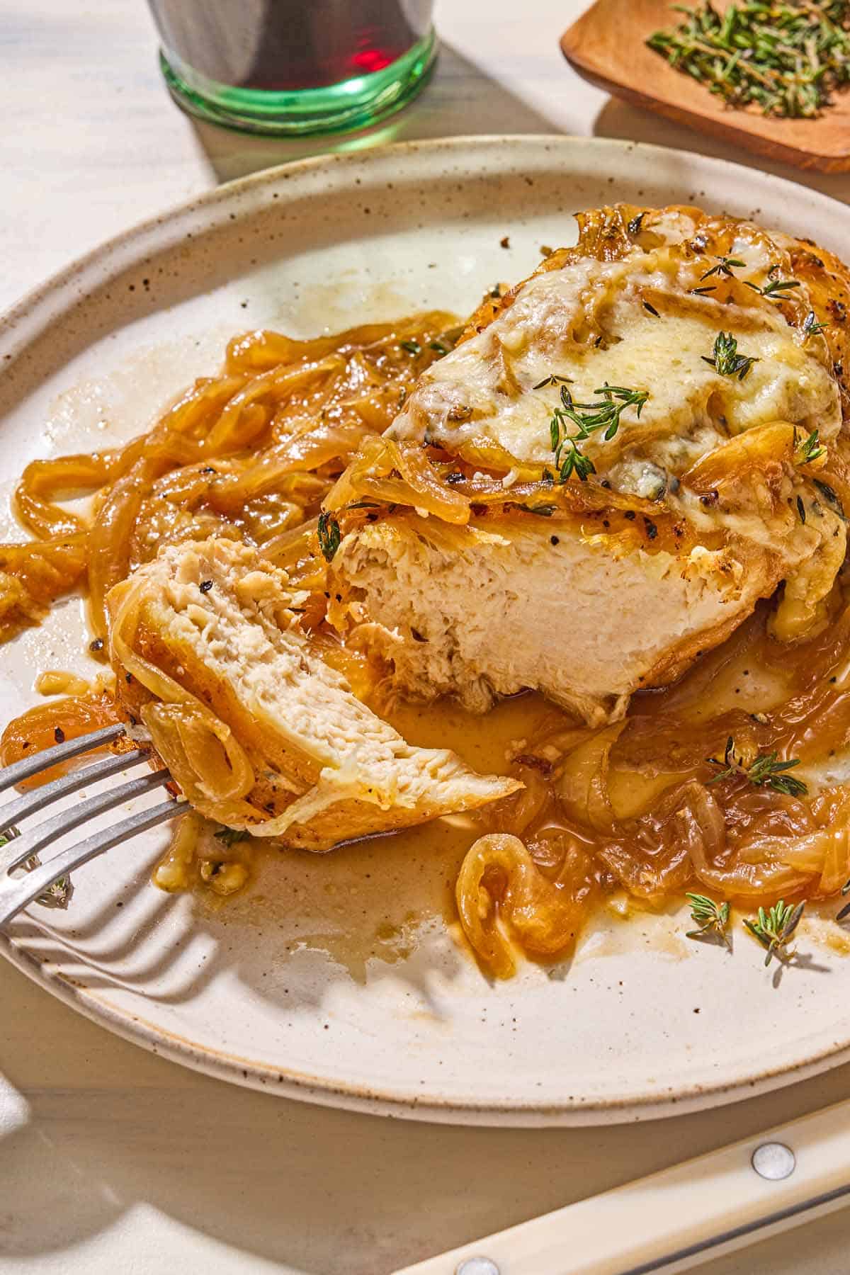 A serving of the French onion chicken on a plate with a fork holding a bite of the chicken. Next to this is a glass of red whine and a bowl of thyme.