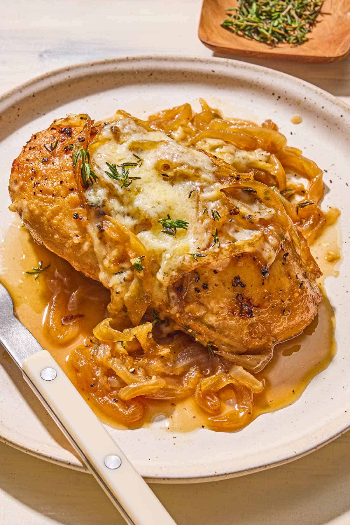 A serving of french onion chicken on a plate with a fork. Next to this is a bowl of chopped thyme.