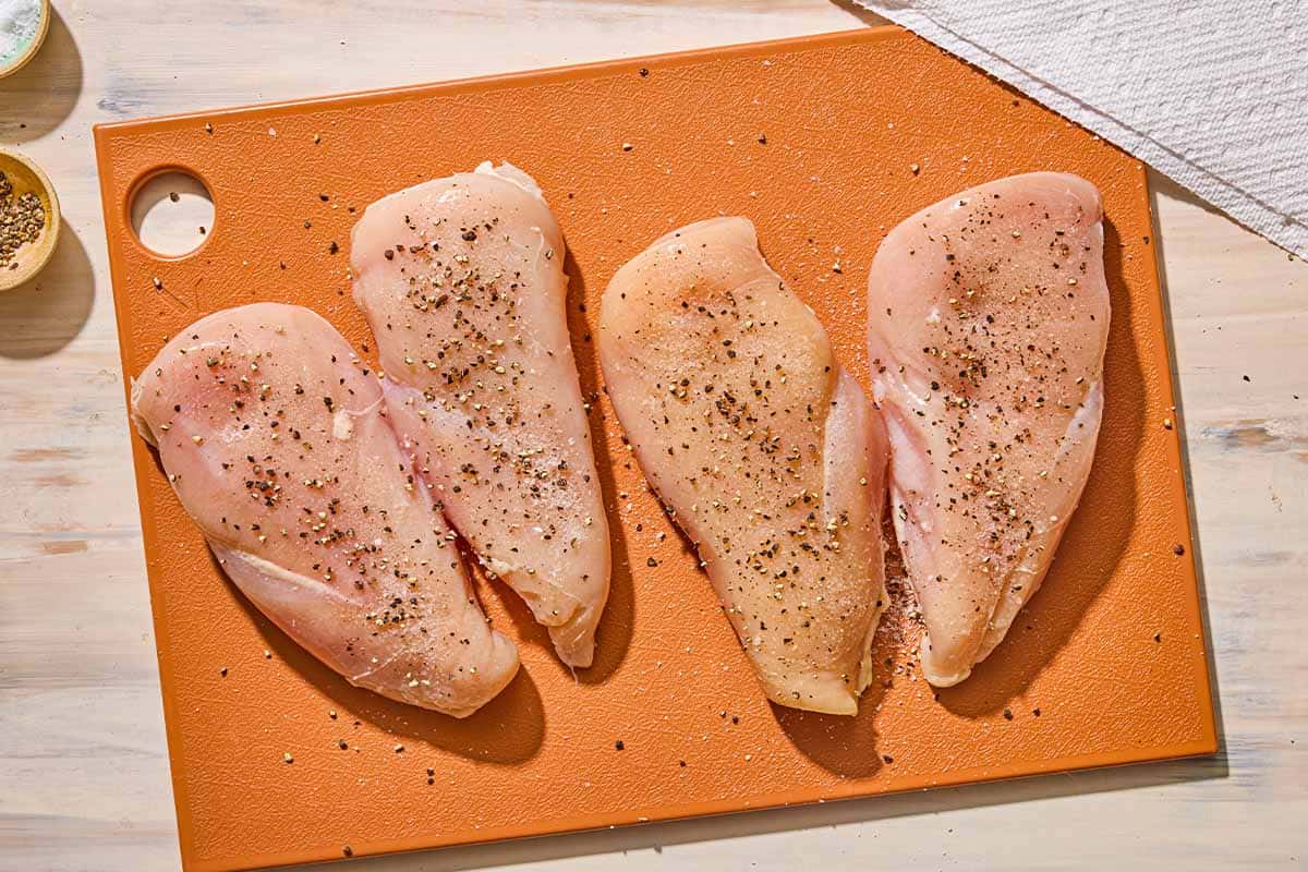 4 raw seasoned chicken breast fillets on a cutting board next to a paper towel and bowls of salt and pepper.