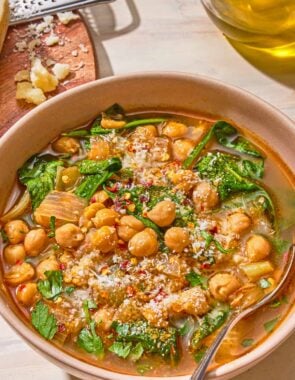 A serving of spinach soup in a bowl with a spoon. Next to this is a block of pecorino Romano cheese, a cheese grater, and a bottle of olive oil.