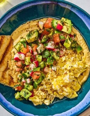 A protein packed breakfast hummus bowl with a pita wedge next to a fork, a spoon and a bowl of za'atar.