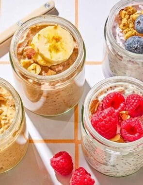 Four jars of overnight oats with various toppings. Next to these are raspberries, blueberries, almonds and a spoon.