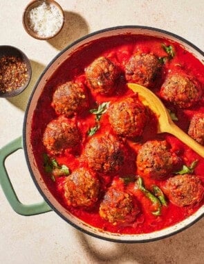 meatballs in sauce in a large pot with a wooden spoon next to small bowls of red pepper flakes and grated parmesan cheese.