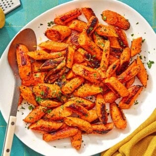 The roasted carrot recipe on a platter with a serving spoon surrounded by a bowl of chopped parsley, a lemon, a bowl of sumac, and a cloth napkin.