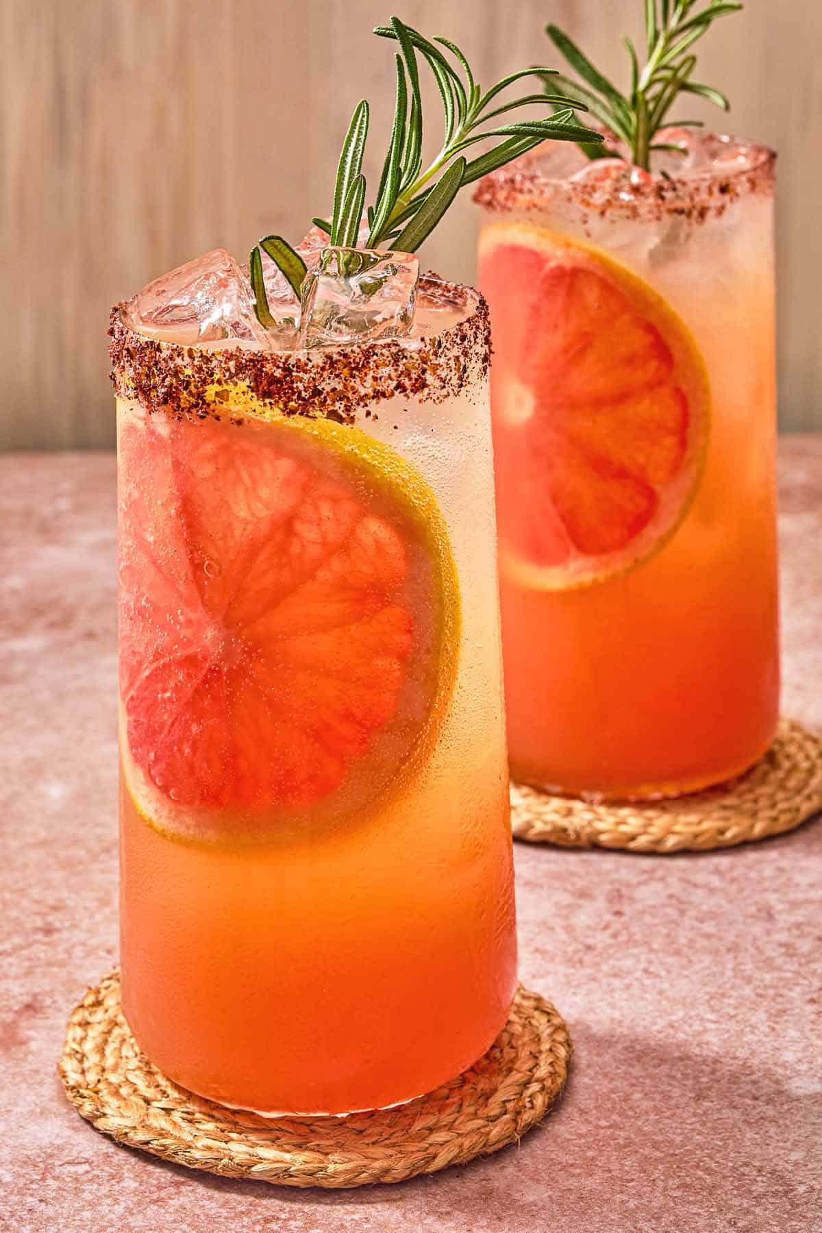 Two glasses of the rosemary grapefruit tonic with grapefruit wheels, sprigs of rosemary and sumac rims on coasters.