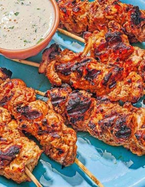 Several shish tawook skewers on a platter with a bowl of tahini sauce.