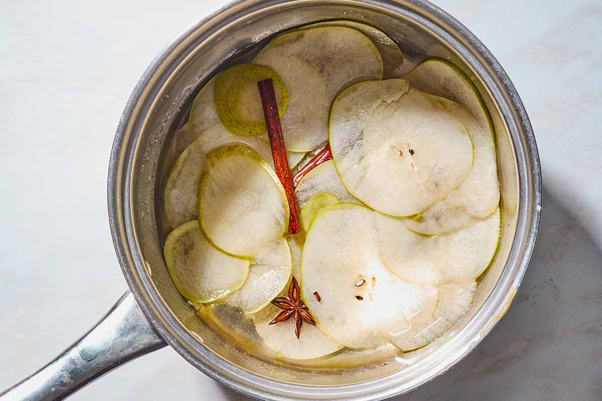 Pear slices simmering in sugar water with star anise and cinnamon sticks in a saucepan.