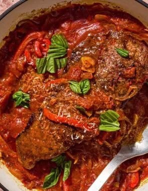 The steak pizzaiola in a skillet with a serving spoon next to parmesan cheese and a cheese grater.