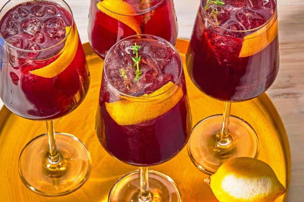 Four tart cherry juice mocktails garnished with lemon twists and sprigs of fresh thyme on a tray with a lemon.