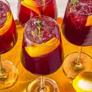 Four tart cherry juice mocktails garnished with lemon twists and sprigs of fresh thyme on a tray with a lemon.