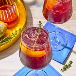Two tart cherry juice mocktails garnished with lemon twists and sprigs of fresh thyme on coasters. Next to this is a tray with the ingredients for the mocktails.