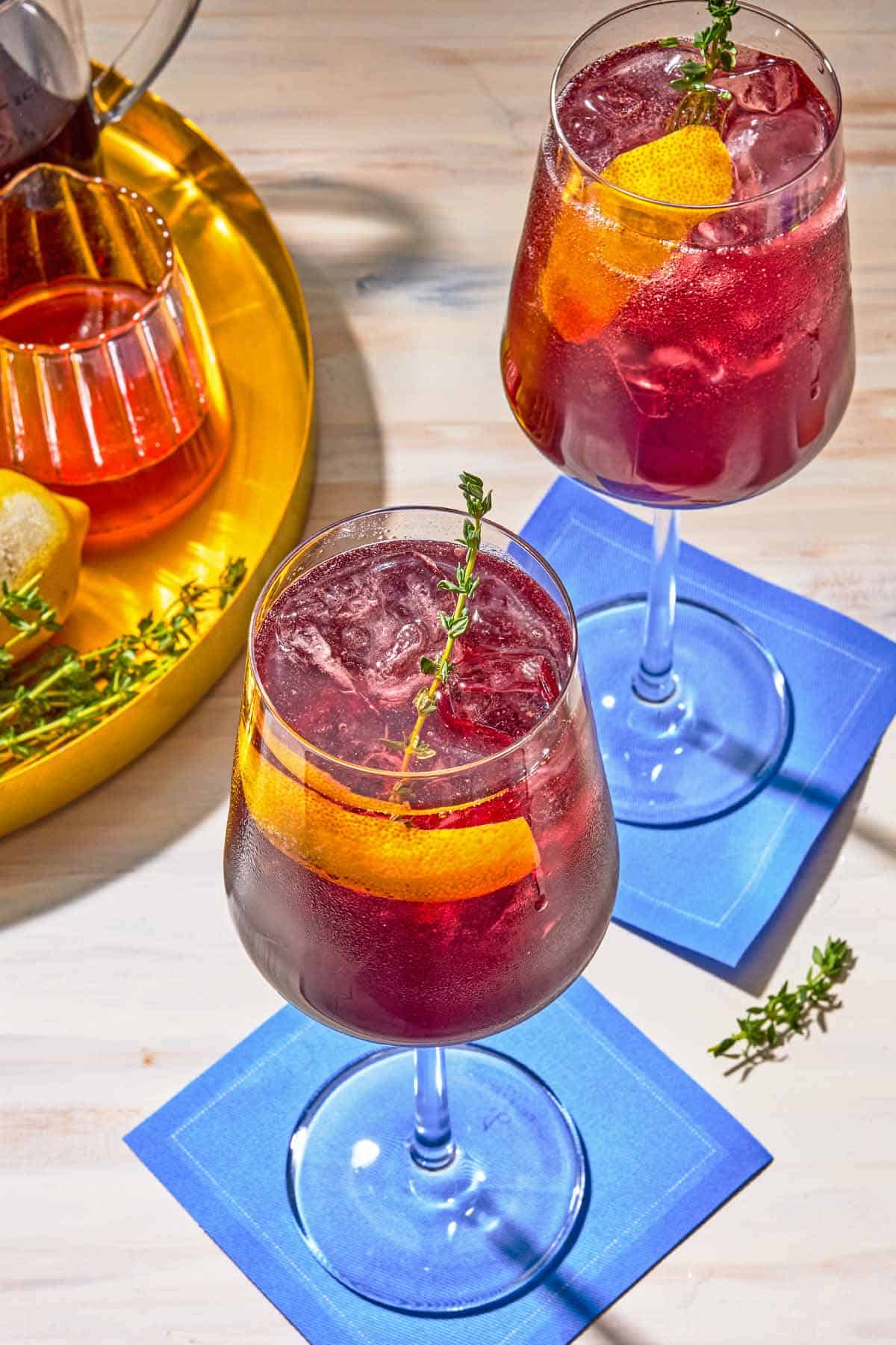 Two tart cherry juice mocktails garnished with lemon twists and sprigs of fresh thyme on coasters. Next to this is a tray with the ingredients for the mocktails.