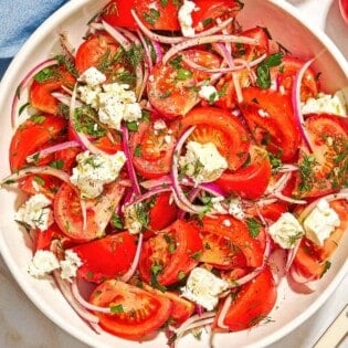 The tomato feta salad in a serving bowl next to a serving spoon, a bowl of sumac, a tomato, and a cloth napkin.