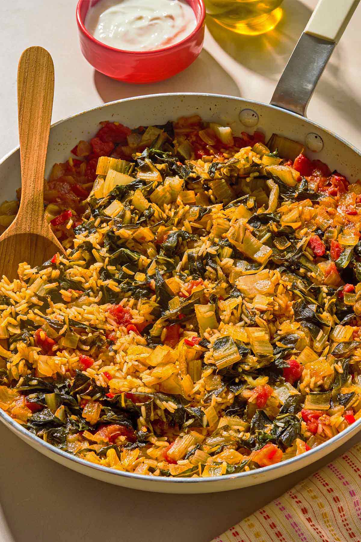 Swiss chard in a skillet with a wooden spoon next to a bowl of yogurt.