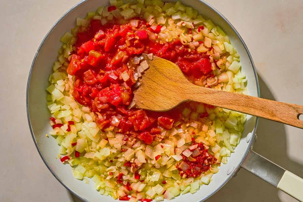 The aromatics for the swiss chard recipe being mixed with the diced tomatoes in a skillet with a wooden spoon.