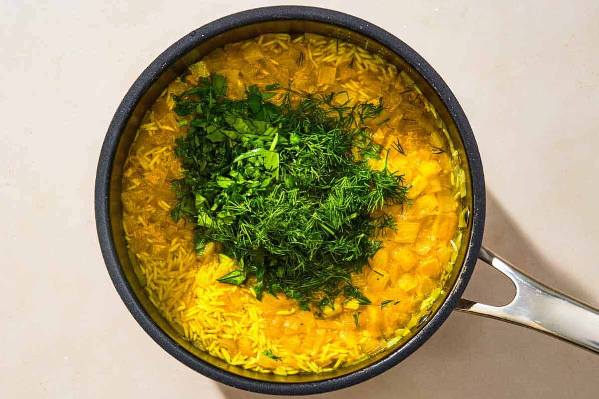 The rice, aromatics, herbs and broth for the turmeric rice in a saucepan.