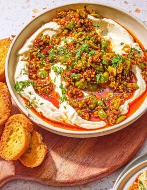Whipped ricotta dip with pistachios, olives and harissa oil in a bowl on a wooden serving board with slices of toasted bread.